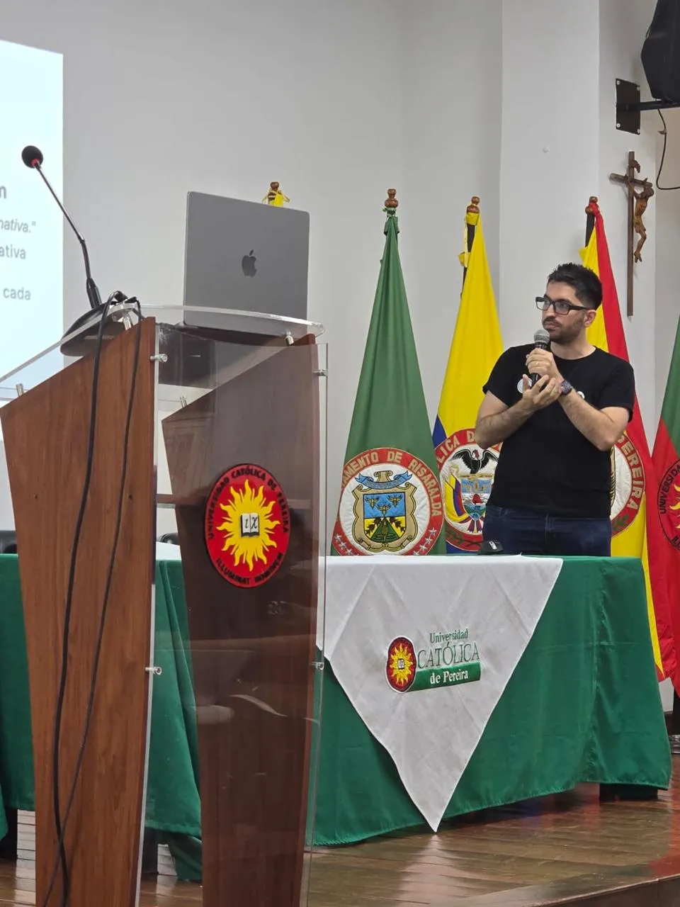 Sergio speaking with a microphone, the Universidad Católica de Pereira table and flags behind him