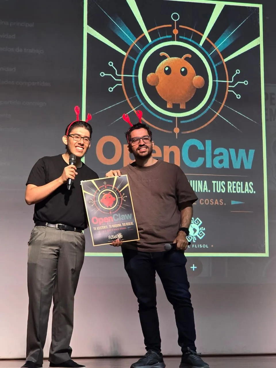 Sergio and a co-host on stage, both wearing red lobster antennae, holding the OpenClaw FLISOL poster in front of the big OpenClaw projection