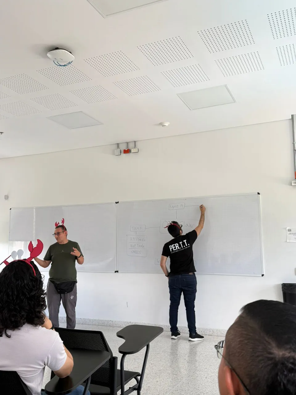 A presenter in a green shirt explaining while another person writes on the OpenClaw diagram, with an attendee in a lobster headband in the foreground
