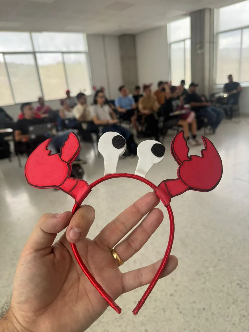 A hand holding up a red lobster-claw headband, the Moltys mascot, with the seated audience blurred behind